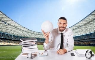 Uomo in abbigliamento formale seduto a una scrivania all’interno di uno stadio di calcio, con libri, pc e pallone, a rappresentare i ruoli manageriali nel calcio e le opportunità per lavorare nello sport.