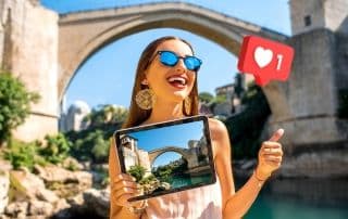 Ragazza con pollice in su davanti a un ponte antico, con un tablet in mano raffigurante il ponte davanti a cui si trova. Affianco a lei c'è un simbolo con cuoricino che rappresenta i like sui social.